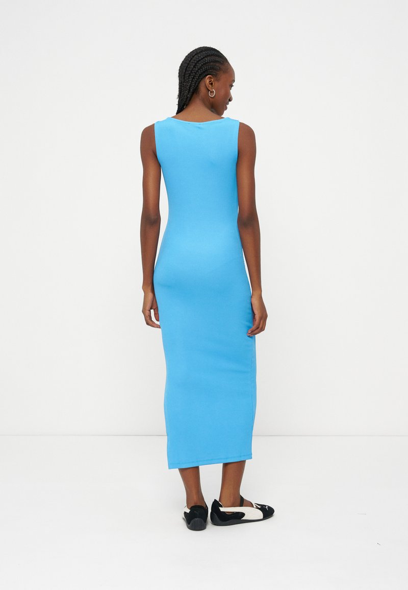 Femme aux cheveux tressés portant une robe midi bleu vif sans manches et des chaussures plates noires et blanches, debout de dos sur un fond blanc.