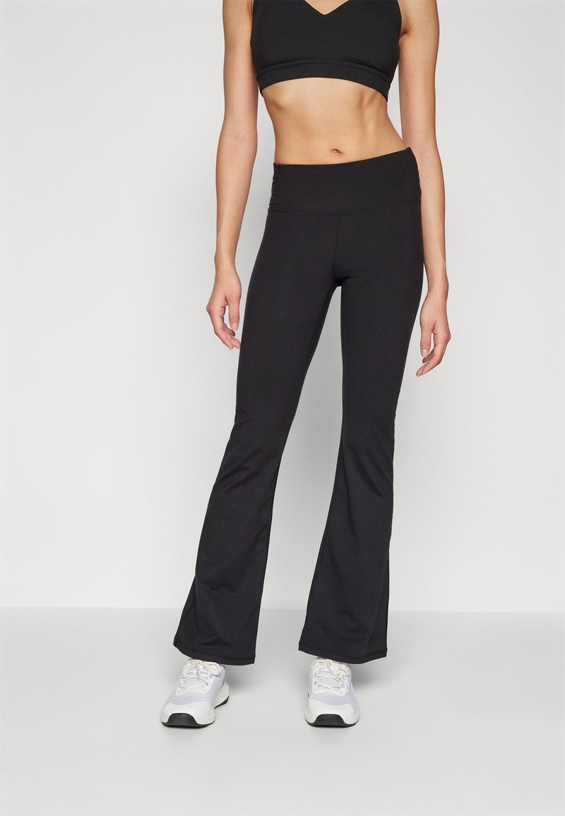 Pantalons de yoga noirs évasés en tissu extensible, dotés d'une taille haute et d'une texture lisse, associés à des chaussures de sport blanches.