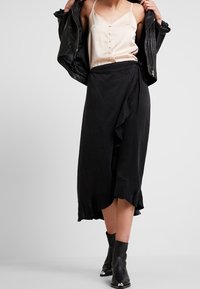 Falda negra de longitud media con dobladillo acampanado, combinada con una blusa de satén beige claro con botones. Lleva puesta una chaqueta de cuero negra y botas de tobillo.