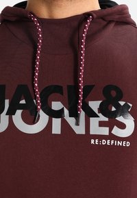 Felpa maroon con cappuccio con grafica testuale in bianco e nero "JACK & JONES RE:DEFINED." Presenta un tessuto testurizzato e cordini con motivo.