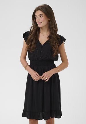 Kobieta o długich brązowych włosach, ubrana w czarną sukienkę z guzikami, z falbanami na rękawach i na dole, stojąca z połączonymi rękoma, lekko się uśmiechając, patrząca w lewo.