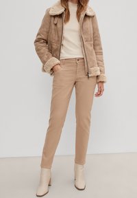 Giacca in pelliccia di pecora marrone con colletto e polsini in pelliccia sintetica, abbinata a pantaloni aderenti beige chiaro e stivali alla caviglia crema. Design semplice e strutturato.