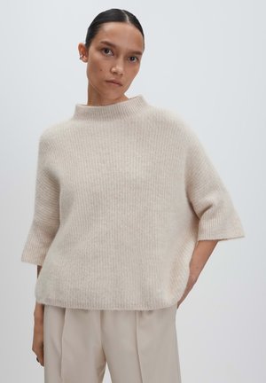 Frau in einem beigen Strickpullover mit weiten Ärmeln und hellbeigen Hosen, stehend vor einem schlichten hellen Hintergrund.