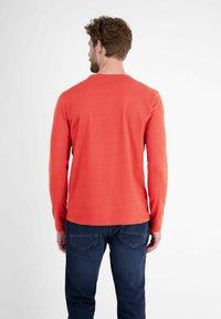 Langärmliges, korallenfarbenes Shirt mit Rundhalsausschnitt. Glatte Textur, einfarbig und figurbetontes Design. Kombiniert mit dunkelblauen Jeans.