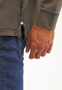 Main droite d'une personne portant une chemise longue à manches brunes et un jean en denim bleu, montrée de la taille jusqu'au milieu de la cuisse sur fond blanc.