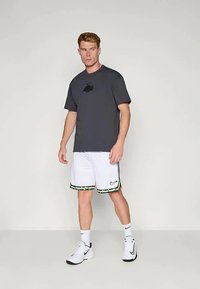 Vīrietis ģērbts tumši pelēkā Nike t-kreklā, baltos Nike šortos ar rakstainu apmalīti, baltās Nike zeķēs un baltās Nike kedās stāv uz pelēka grīdas.