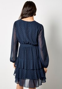 Granatowa sukienka z długimi przezroczystymi rękawami, warstwową falbaną u dołu i wąską talią, zdobiona małymi, białymi kropeczkami.