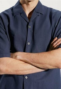 Granatowa koszula z krótkim rękawem z lekkiego materiału, wyposażona w kołnierzyk typu camp oraz dwa guziki z przodu. Gładka tekstura i luźny krój.