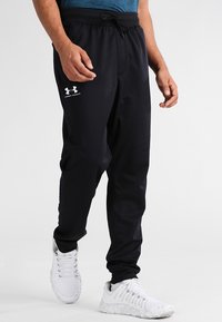 Mann trägt schwarze Under Armour Jogginghosen und weiße Sportschuhe, steht vor einem schlichten hellen Hintergrund.