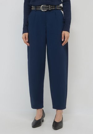 Eine Frau trägt marineblaue, schmal geschnittene Hosen, schwarze High Heels und einen schwarzen Gürtel mit silbernen Ösen und Schnalle und steht vor einem schlichten, hellen Hintergrund.