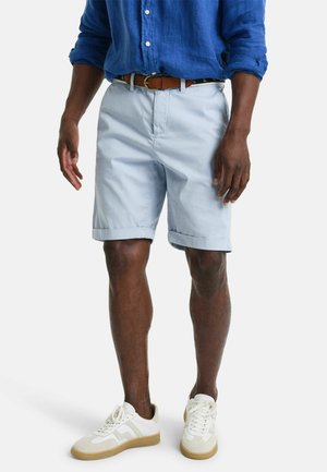 Shorts di cotone azzurro con risvoltini, dotati di tasche laterali e chiusura con bottone, indossati con una camicia di lino blu e sneakers bianche.