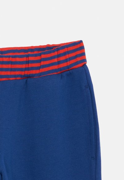 Calças azuis com uma cintura elástica riscada em vermelho e azul e um bolso lateral em fundo branco.