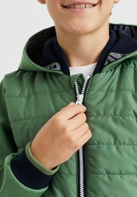 Grön vadderad jacka med dragkedja fram, kontrasterande marinblå huva och muddar, samt quiltat mönster. Har en vit dragkedja och logotypdetalj invändigt.