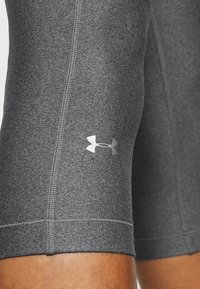 Szare sportowe spodenki o gładkiej fakturze, z wyraźnym szwem i logo Under Armour w srebrnym kolorze na lewej nogawce.