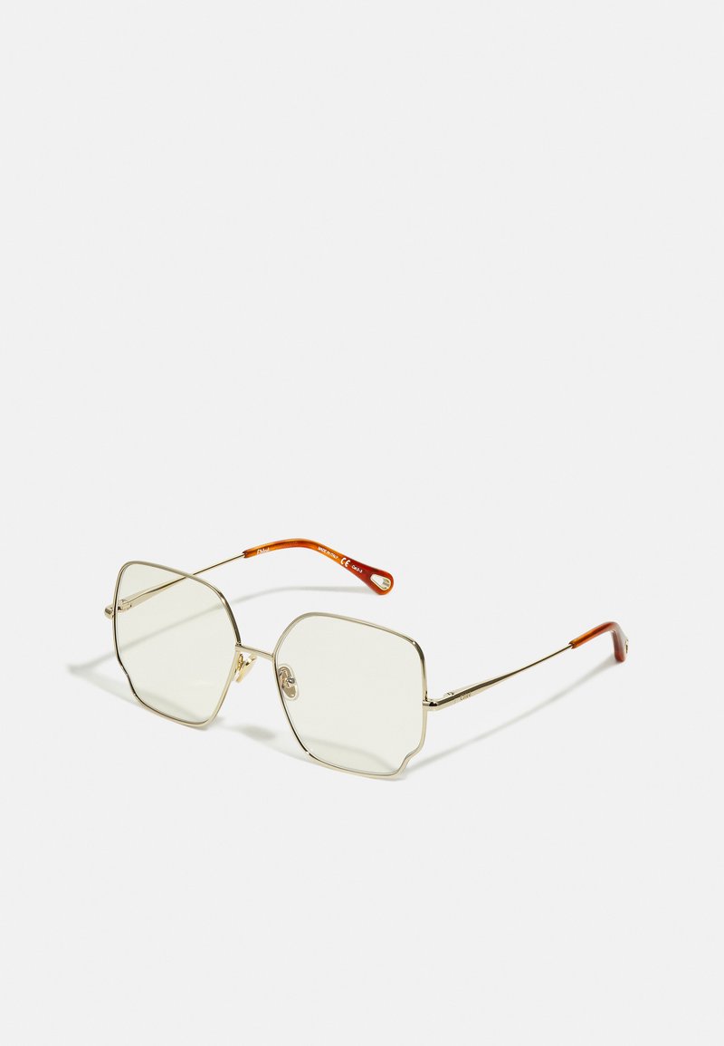 Lunettes en métal doré avec une forme octogonale unique, dotées de lentilles transparentes et de pointes de branches accentuées en orange. Design minimaliste.