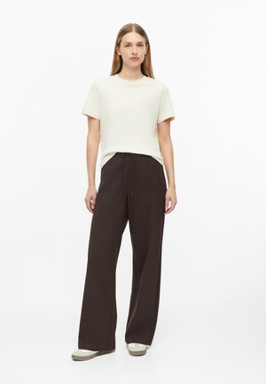 Femme debout portant un haut crème à manches courtes, un pantalon large marron foncé avec cordon de serrage, et des chaussures blanches sur un fond blanc.