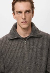 Suéter gris con cremallera, textura acanalada y cuello amplio. La tela parece cálida y gruesa, con un patrón jaspeado sutil.