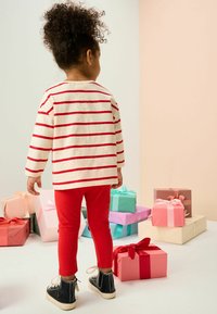 Maglietta a maniche lunghe a righe rosse, leggings rossi e scarpe alte nere. Bambino di spalle, circondato da diverse scatole regalo dai colori pastello.