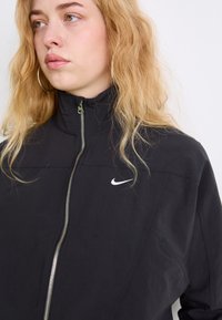 Juodas užtrauktuku užsegamas džemperis su aukštu apykakle, sidabriniu užtrauktuku ir mažuoju baltuoju Nike logotipu ant krūtinės. Pagamintas iš lygaus, lengvo audinio.