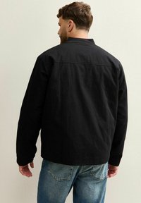 Man in een zwarte lange mouwen jas en blauwe spijkerbroek die met zijn rug naar een effen beige muur staat.