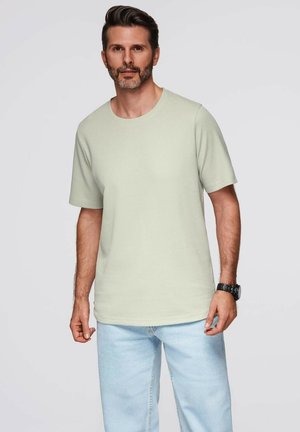 Mand med kort brunt hår og skæg iført en beige kortærmet skjorte og lyseblå jeans, der står mod en ensfarvet baggrund.