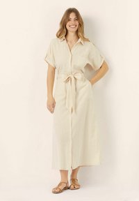 Vestito camicia in misto lino beige con maniche corte arrotolate, bottoni frontali, una cintura in vita e una silhouette fluida, abbinato a sandali brown con fasce.