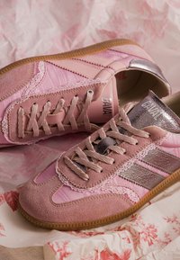 Paire de baskets roses avec des accents en daim et métal, design à lacets et semelles en caoutchouc à motifs sur fond floral et papier froissé.