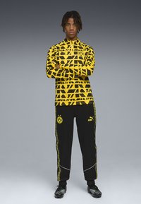 Sportjacke und Hose Set, gelb mit schwarz geometrischem Muster, ausgestattet mit Teamlogo. Schwarze Hose mit gelben Akzenten und Logo am Bein.