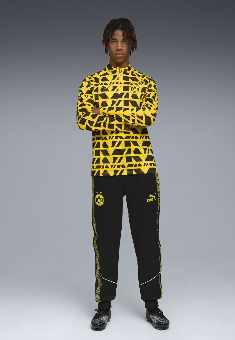 Set giacca sportiva e pantaloni, giallo con motivo geometrico nero, con logo della squadra. Pantaloni neri con accenti gialli e logo sulla gamba.