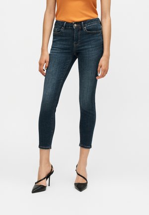 Kvinde iført mørkeblå skinny jeans, orange ærmeløs top og sorte højhælede sko, står mod en ensfarvet baggrund.