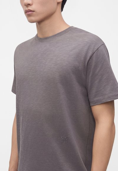 Jeune personne portant un t-shirt gris uni à manches courtes avec un petit logo circulaire cousu près de la taille sur fond blanc.