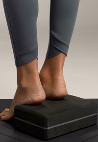 Bloc de yoga negru cu o formă dreptunghiulară în relief, sprijinind tălpile goale. Colanții gri au un design ajustat cu un detaliu subtil de cusătură.