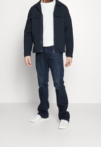 Chaqueta navy con dos bolsillos en el pecho, camiseta blanca, jeans oscuros y zapatillas blancas. La chaqueta tiene cierre de cremallera y un corte relajado.