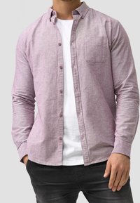 Lys lilla skjorte med knapper, lavet af struktureret stof, med en brystlomme, standart krave og båret over en hvid T-shirt.