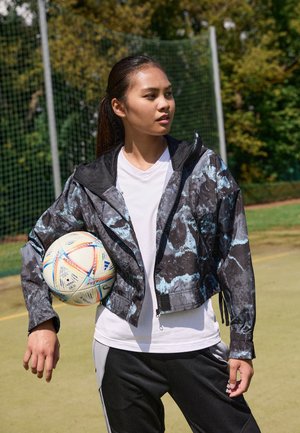 Cropped black jacket with blue abstract patterns, mesh-lined hood, zip closure, paired with a white t-shirt and black athletic pants, holding a soccer ball.