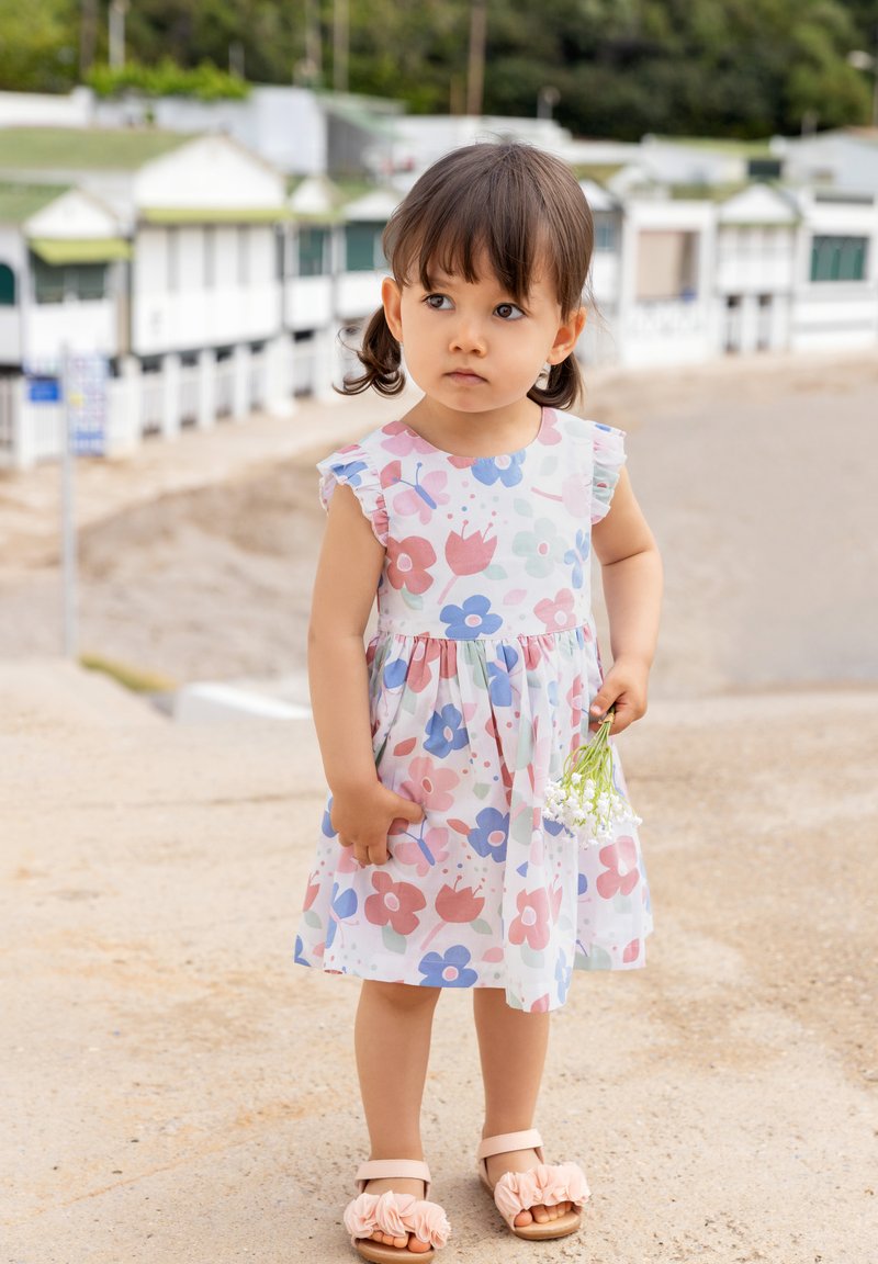 Robe en coton à motifs floraux avec manches courtes à volants, mettant en vedette des fleurs roses, bleues et rouges. Associée à des sandales ornées de volants en tissu.