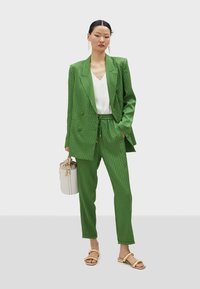 Green patterned blazer and trousers set with a white top. Model holds a cream handbag; wearing light golden sandals. Satin texture.