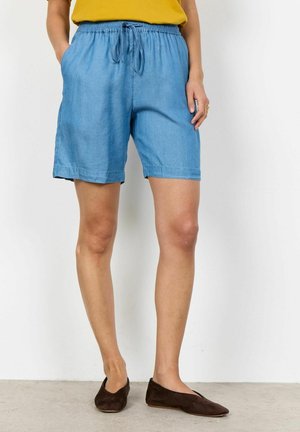 Person trägt hellblaue Shorts mit Kordelzug, senfgelben Oberteil und dunkelbraune flache Schuhe, steht vor weißem Hintergrund.