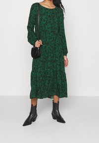 Vestido verde de manga larga con un diseño de estampado floral, cintura fruncida, falda en capas, combinado con botas negras hasta el tobillo y un bolso pequeño negro.