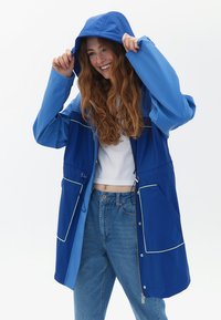 Giacca impermeabile blu con cappuccio, chiusura con zip e pannello azzurro a contrasto. Presenta ampie tasche frontal e cordoncino in vita.