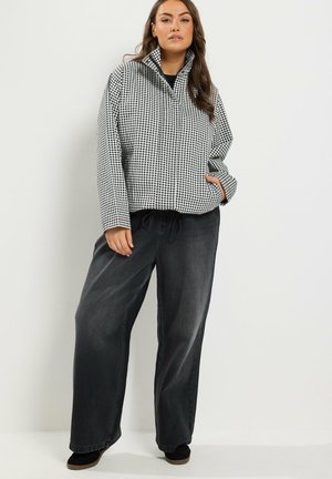 Donna con capelli castani lunghi che indossa una giacca a quadri bianca e nera e pantaloni neri larghi, in piedi davanti a uno sfondo chiaro e uniforme.