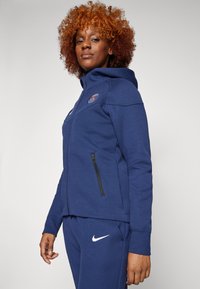 Navyfarbener Reißverschluss-Hoodie mit einem Logo auf der Brust und schwarzen Reißverschlussdetails, kombiniert mit passender Jogginghose mit einem weißen Nike-Logo.