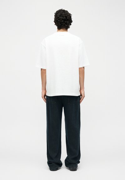 Άνδρας που φοράει απλό λευκό φαρδύ κοντομάνικο μπλουζάκι και άνετα μαύρα τζιν, στεκόμενος με την πλάτη προς τα εμάς σε λευκό φόντο.