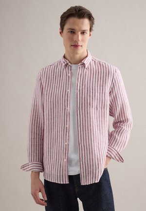 Jonge man die een wit T-shirt en een rood-wit gestreept overhemd met knopen draagt, staand met één hand in zijn zak.