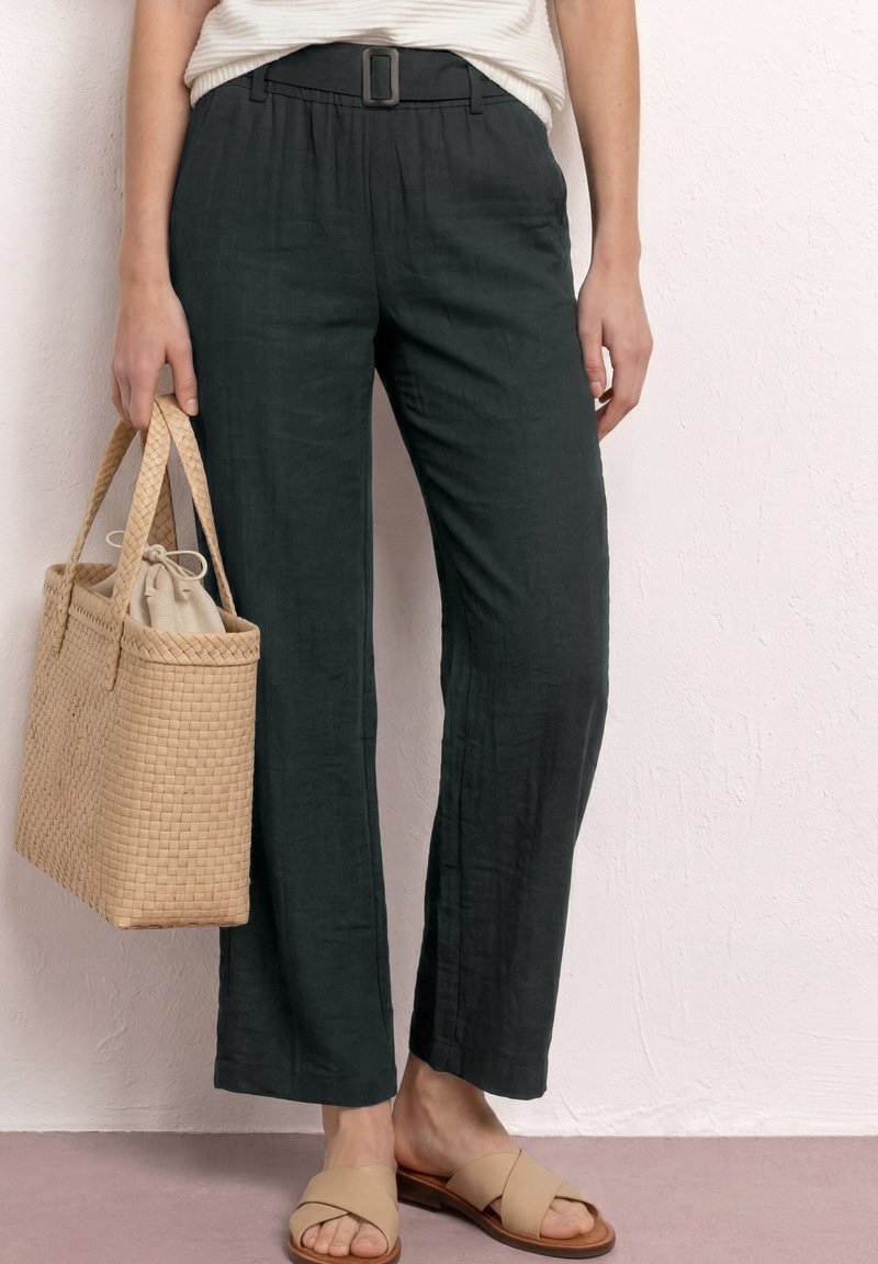 Vrouw draagt zwarte broek met hoge taille en riem, beige sandalen met kruisbandjes, houdt een geweven beige tote bag met stoffen inzetstuk vast.