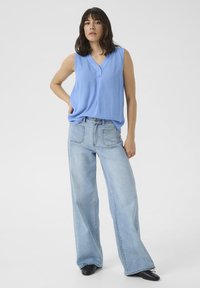 Blouse sans manches bleu clair avec un décolleté en V et des détails de boutons, associée à un jean en denim clair à taille haute et jambes larges. Chaussures noires.