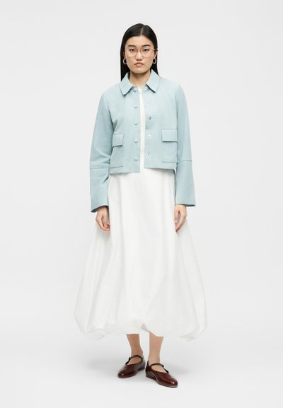 Γυναίκα που φοράει ανοιχτό γαλάζιο κοντό μπουφάν πάνω από λευκό φόρεμα μέχρι τους αστραγάλους, στρογγυλά γυαλιά, σκουλαρίκια κρίκους και σκούρα κόκκινα παπούτσια Mary Jane, όρθια.