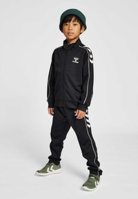 Chándal negro con acentos blancos y logo, combinado con zapatillas verdes. Cuenta con una chaqueta con cremallera y pantalones con cintura elástica.