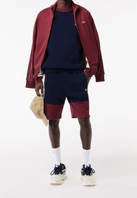 Chaqueta granate con cremallera sobre sudadera azul marino, combinada con pantalones cortos azul marino y granate. Calcetines blancos y gorra beige completan el look deportivo.