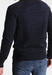 Granatowy sweter dzianinowy z teksturowanymi paskami, okrągłym dekoltem oraz ściągaczami przy mankietach i u dołu. W połączeniu z jasnoniebieskimi dżinsami.